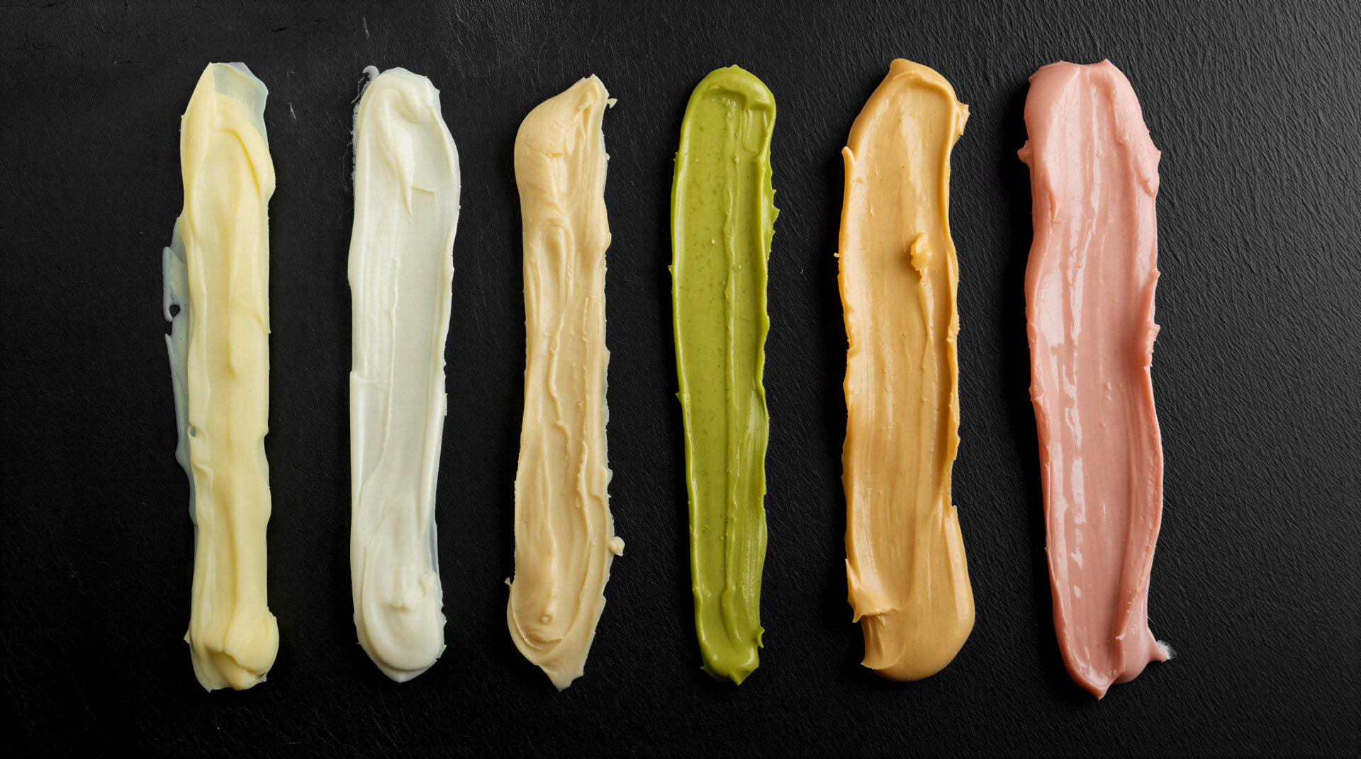 Sechs Streifen verschiedener Aufstriche wie Butter, Mayonnaise, Remoulade und Guacamole nebeneinander auf schwarzer Steinplatte